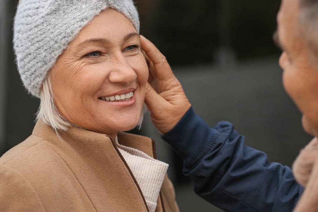 Une femme âgée au bonnet gris sourit, une main posée sur la joue.